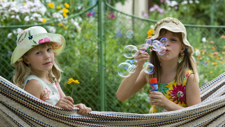 Two girls in summer dresses and hats are sitting in a hammock. One is blowing bubbles, the other is holding a yellow flower.
