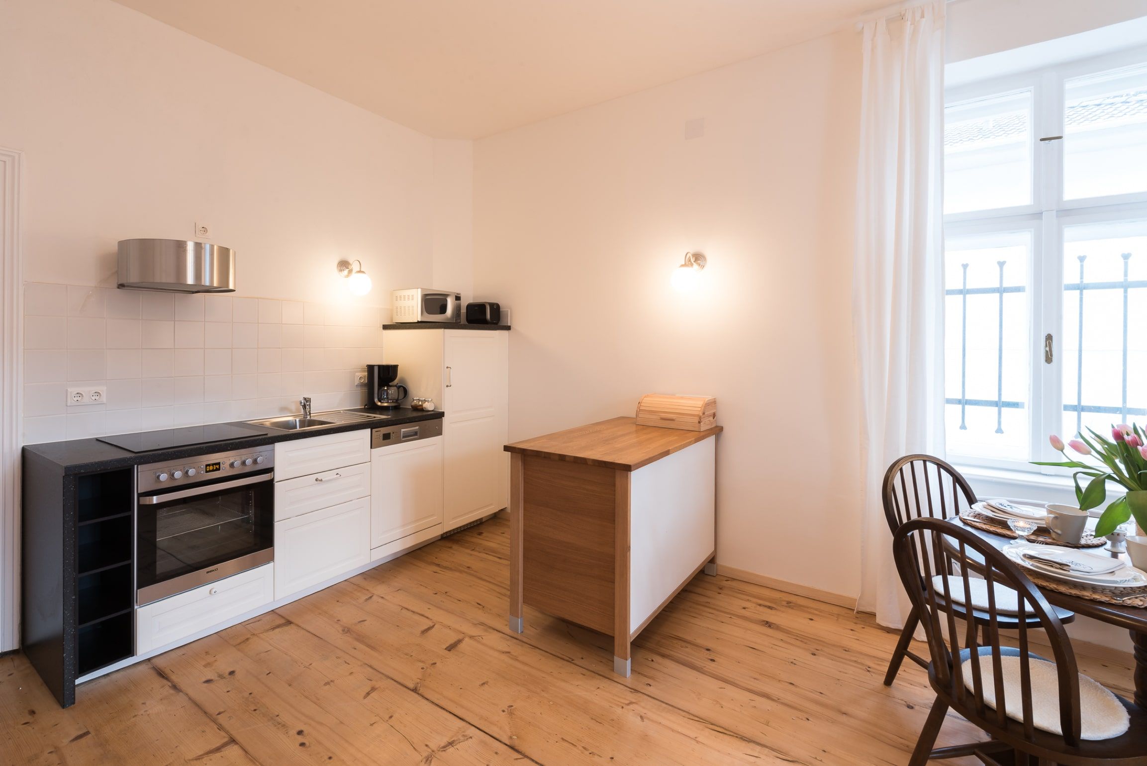Modern kitchen with wooden floor, white furnishings and small dining table by the window.