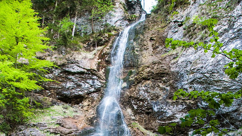 A waterfall flows over a rocky cliff, surrounded by green vegetation.