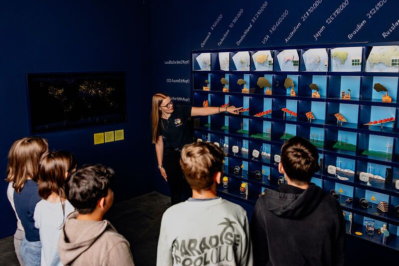 A group of young people watch a woman pointing at a wall of diagrams and models during a guided tour.