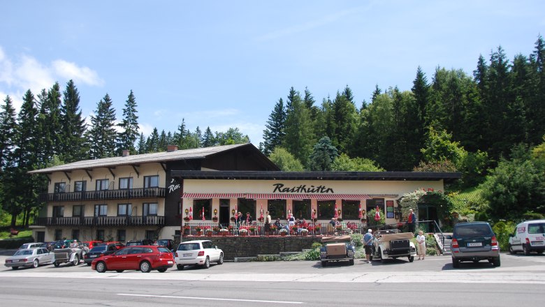 A rest house with a terrace and parked cars in front of it, surrounded by trees.