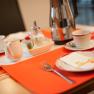 Breakfast table with cups, plates, cutlery and coffee pot on an orange place mat.