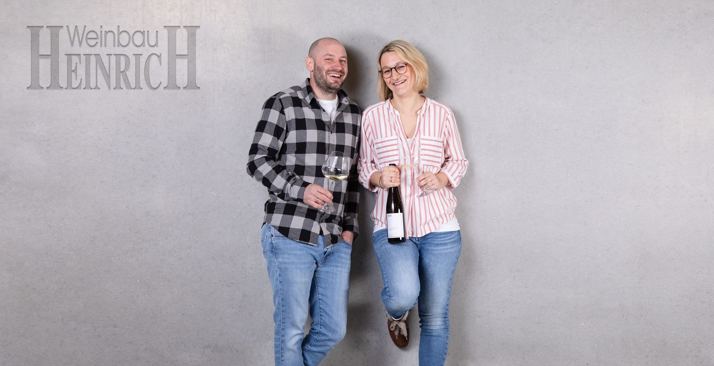 Two people stand smiling in front of a gray wall with wine glasses in their hands. On the left is a man, on the right a woman. At the top left it says 'Weinbau Heinrich'.