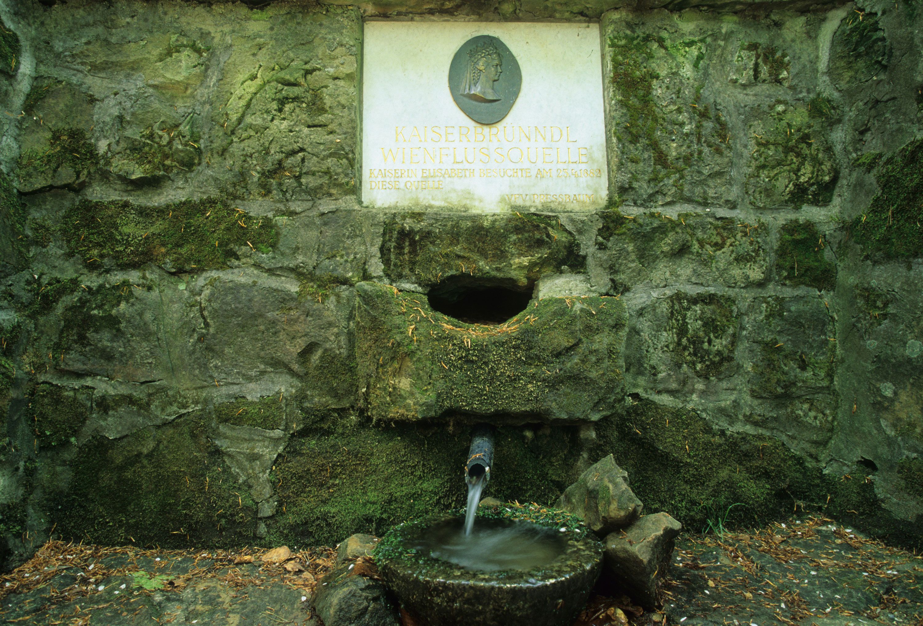 Stone fountain with inscription 'Kaiserbründl Wienflussquelle'. Water flows from a pipe into a bowl.