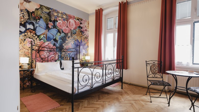 A stylishly furnished room with a wrought-iron bed, floral wallpaper and a small table with chairs in front of large windows.