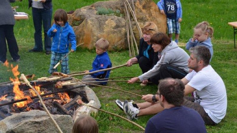 People sit around a campfire and roast bread on long sticks.