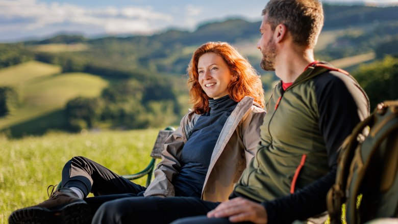 Two people are sitting on a bench in a green landscape.