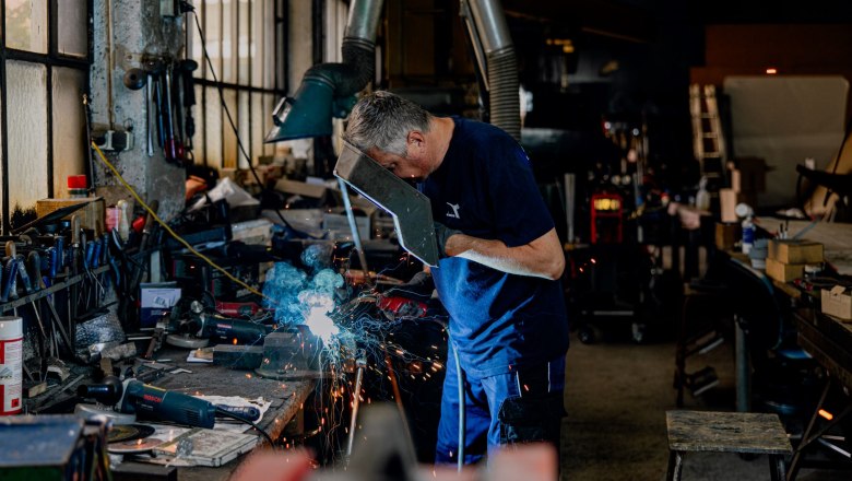 A man is welding in a workshop, surrounded by tools and sparks.