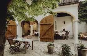 Cozy guest garden with wooden tables and chairs under a tree, surrounded by white walls and open wooden doors.