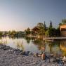 Wooden chalets on the shore of a small lake with a jetty and plants in the foreground.