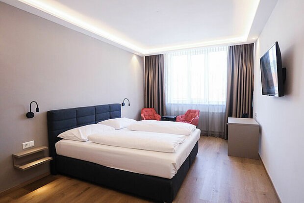 Modern hotel room with a double bed, two red armchairs, a large window and a flat-screen TV on the wall.