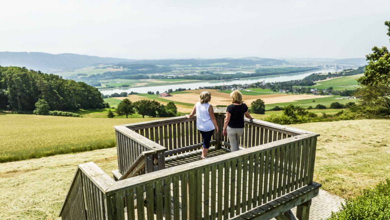 Two people stand on a wooden viewing platform with a view of a wide landscape and a river.