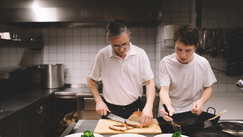 An older man and a younger man work together in a kitchen. The older man cuts meat while the younger man puts broccoli on a plate.
