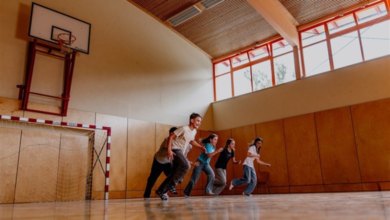 Five teenagers running in a gym with a basketball hoop and handball goal.