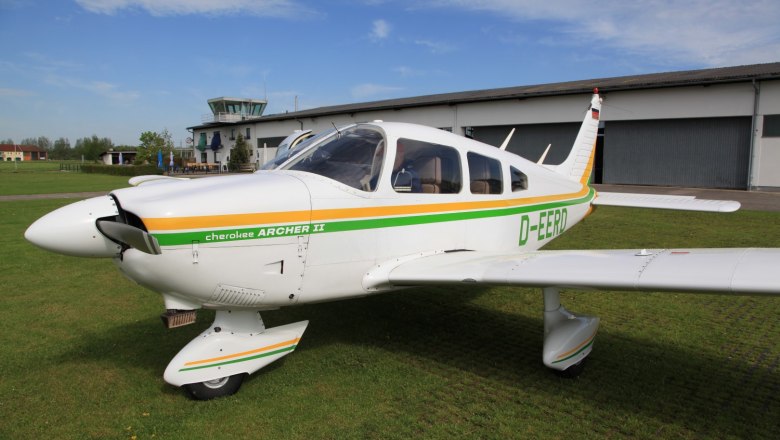 A small airplane, Piper Cherokee Archer II, stands on a green field in front of a hangar and a control tower.