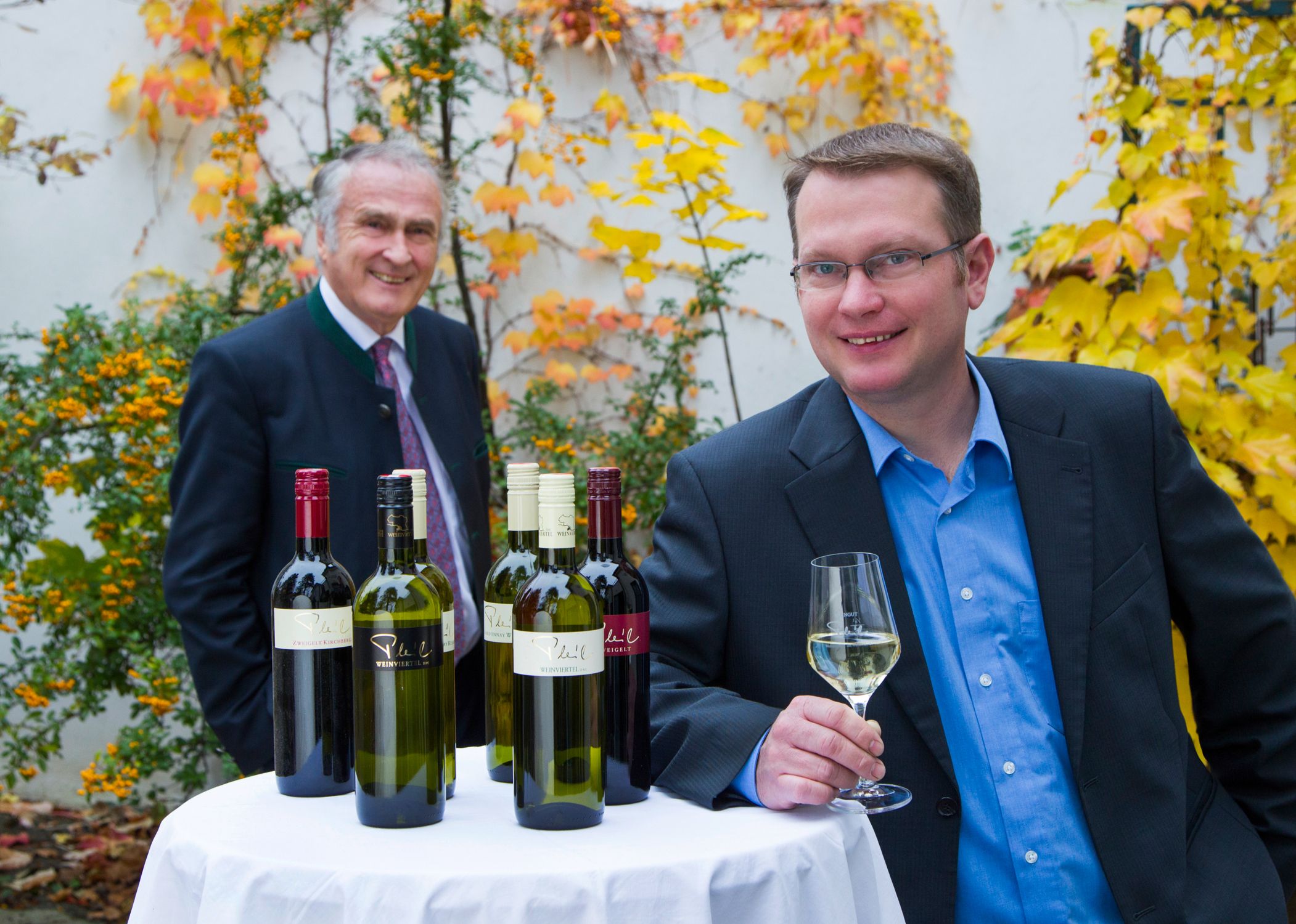 Two men stand at an outdoor table with bottles of wine, surrounded by autumnal plants.