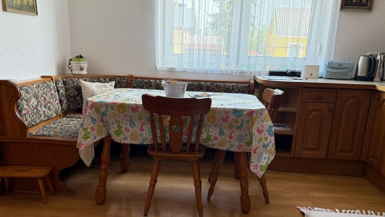 Dining area with wooden table, chairs and corner bench, colorful tablecloth, window in the background.