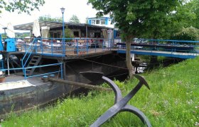 Exterior view of a restaurant on an old ferryboat with blue balustrade and green surroundings.