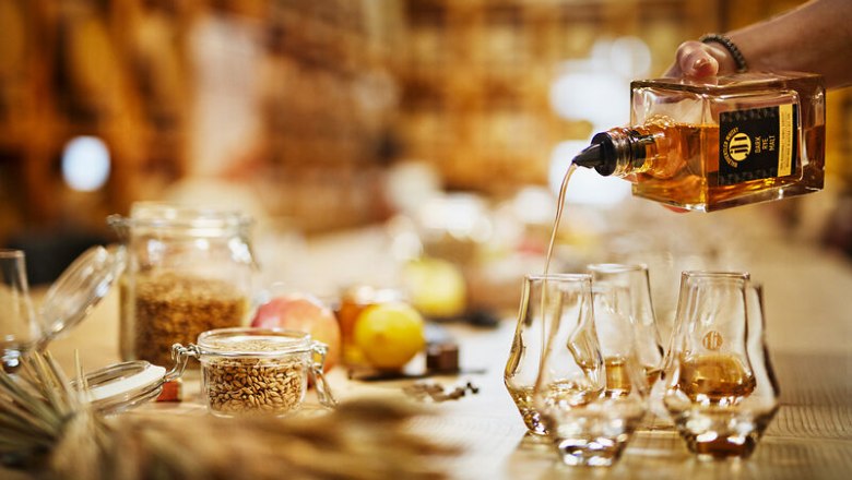 A hand pours whisky from a bottle into several glasses on a table with cereals and fruit.