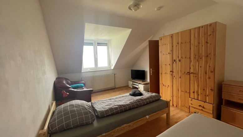 A simply furnished bedroom with two beds, a closet, a TV and an armchair under a sloping roof.