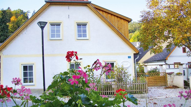 A white cottage with red roof tiles and a garden fence, surrounded by blossoming flowers and trees in the fall.
