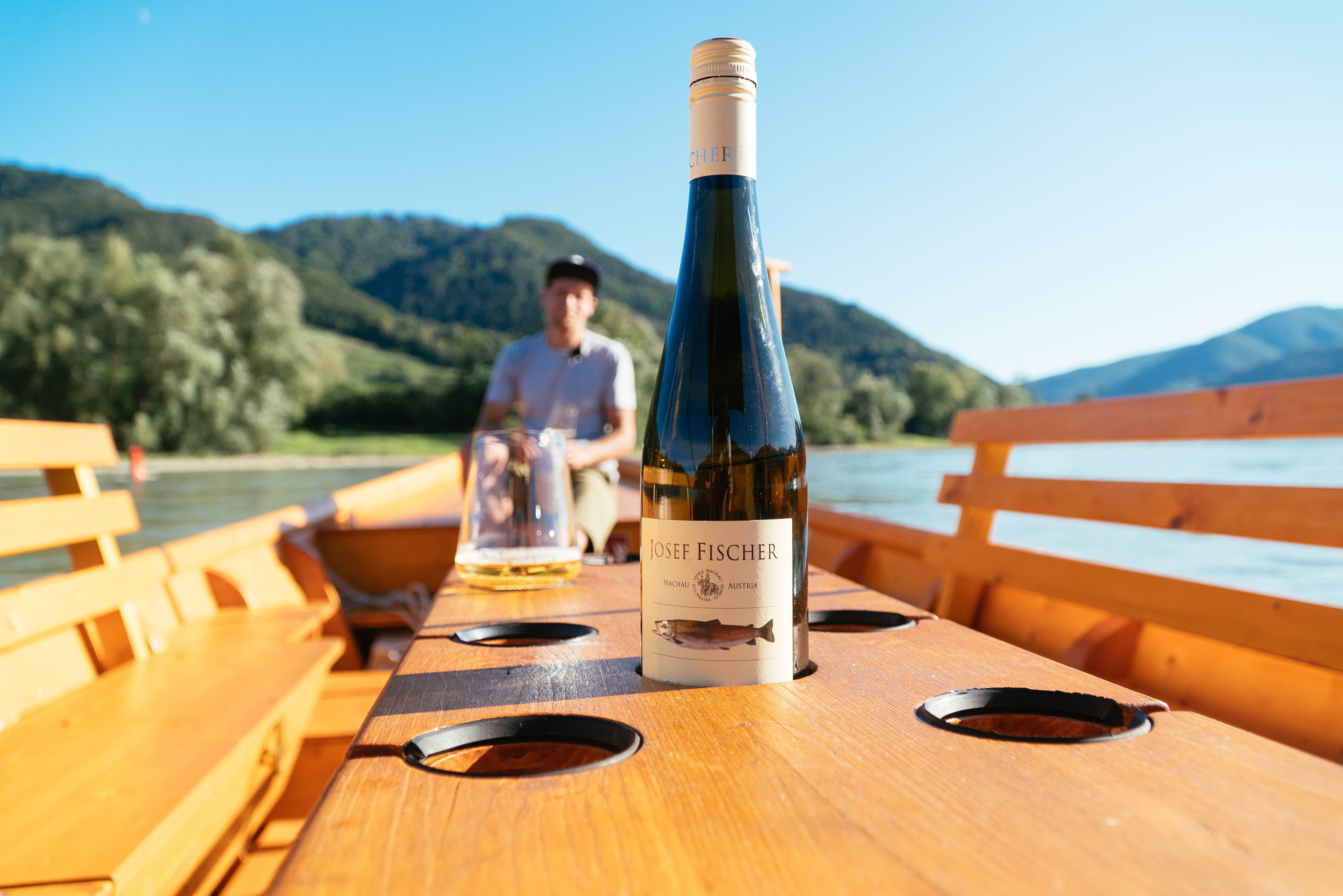 Wine bottle on a boat table with blurred background of mountains and a man.