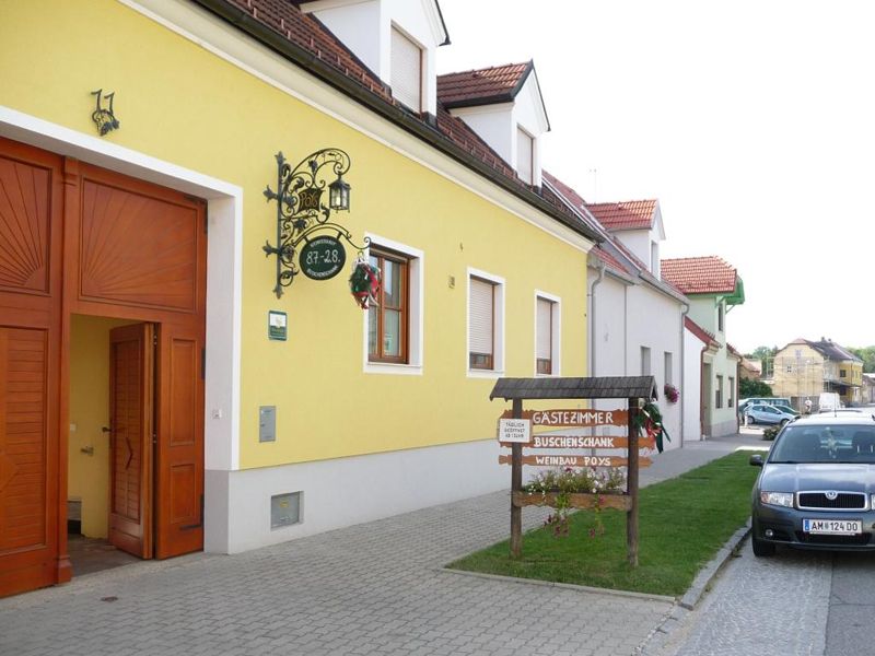 Yellow building with sign 'Gästezimmer Buschenschank Weinbau Poys' and parked car in front of it.