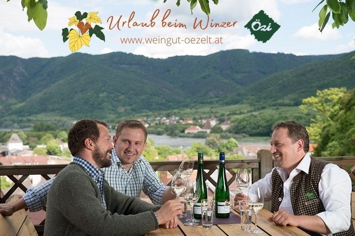 Three men sit at a table with bottles of wine and glasses, a picturesque landscape with hills in the background.