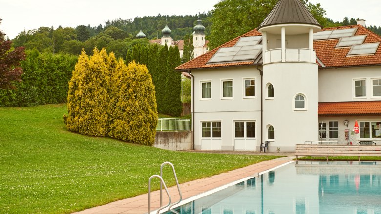 Exterior view of a leisure facility with pool and building, trees and church spires in the background.