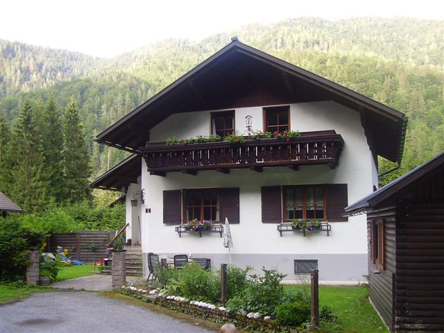 A traditional vacation home with a wooden balcony and flower boxes, surrounded by green trees and mountains.