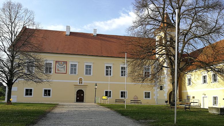 Yellow castle building with red roof and tower, surrounded by trees.