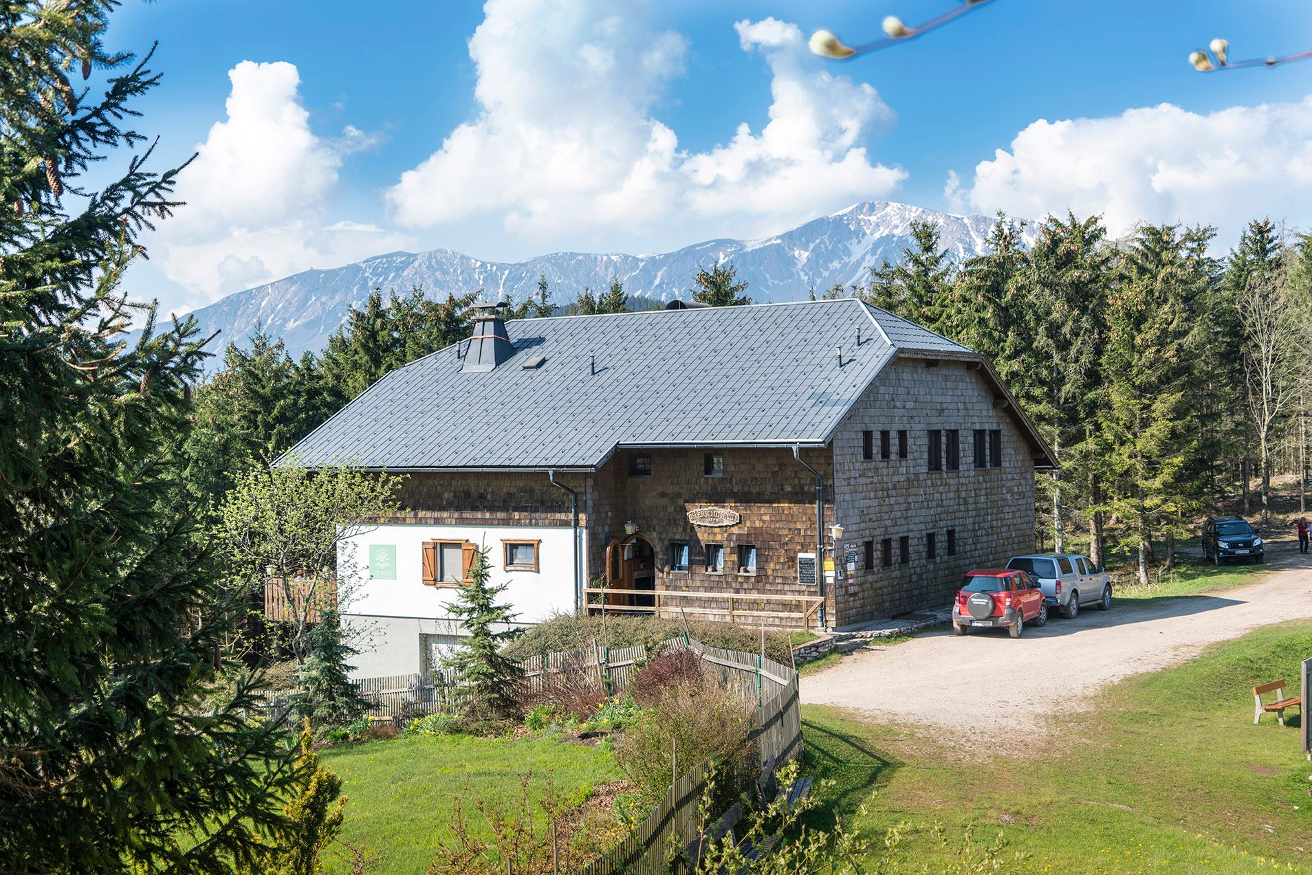 A rustic shelter in a mountainous landscape with trees and cars in the foreground.