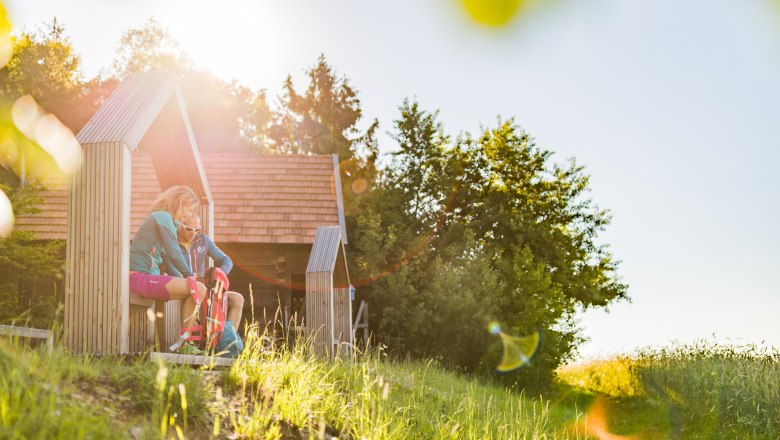 Two people sit in a wooden structure in a sunny landscape.