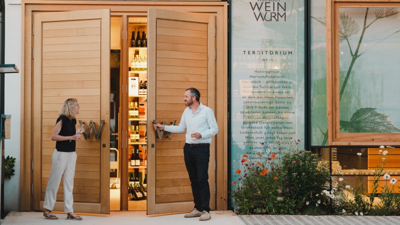 Two people are standing in front of a winery with open wooden doors showing bottles inside.