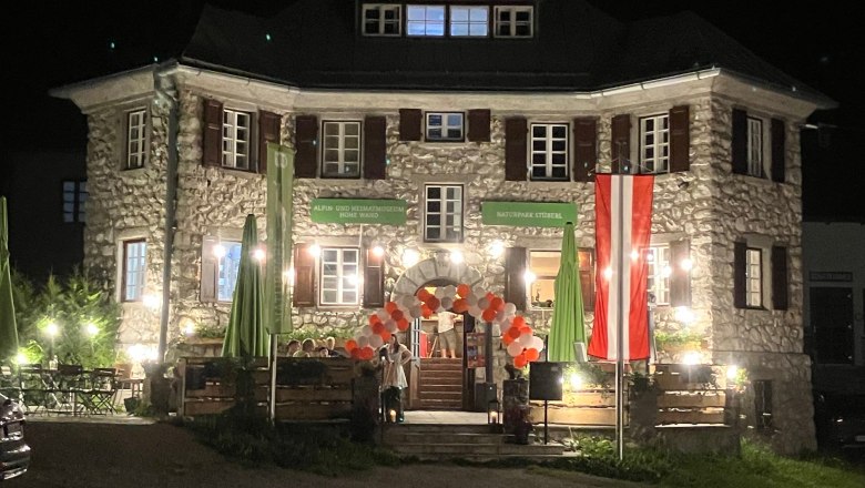 An illuminated stone building at night with an Austrian flag and balloons at the entrance.