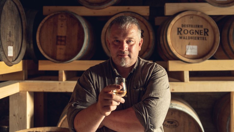 Man in a distillery with a glass in his hand in front of wooden barrels.