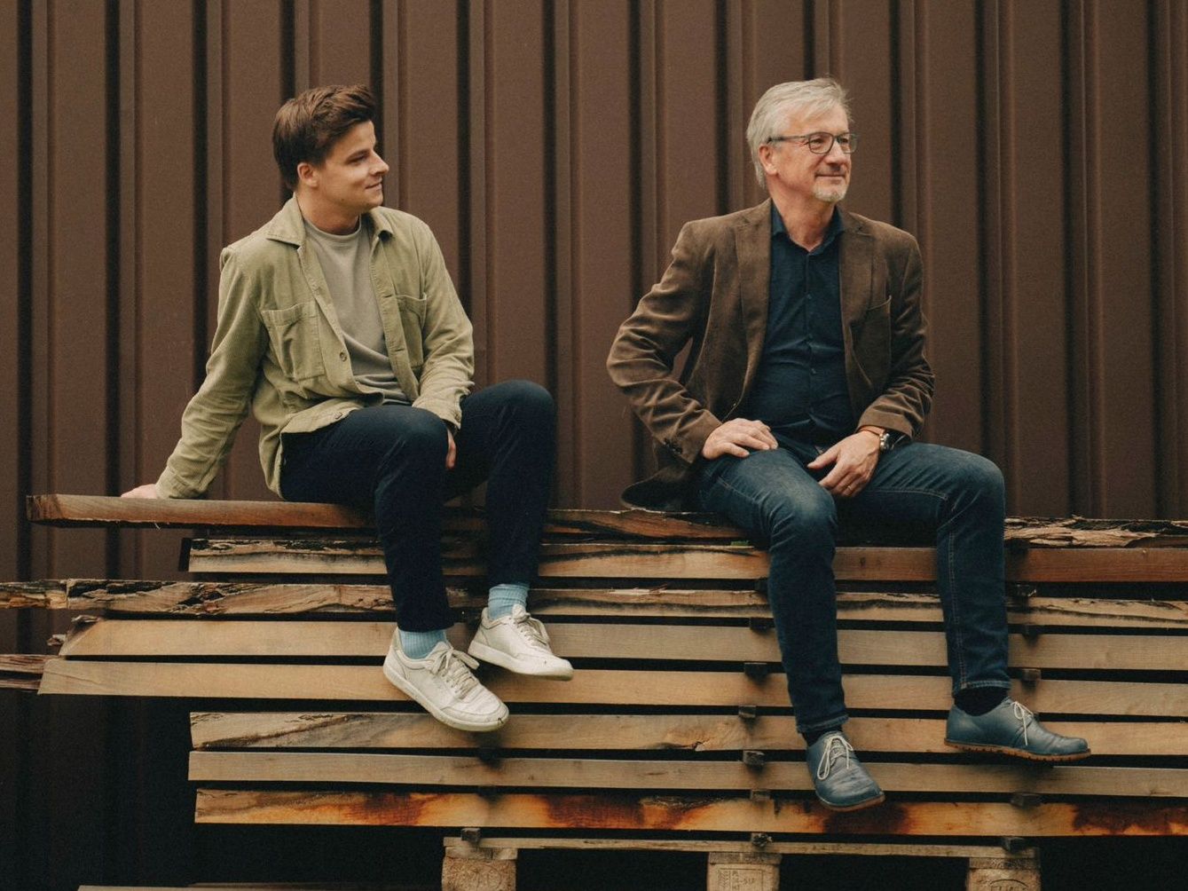 Two men sit relaxed on stacked wooden planks in front of a dark wooden façade and look to the side; a calm, natural atmosphere in warm colors.