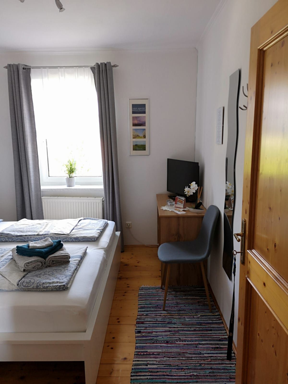 Bright bedroom with double bed, gray curtains, small desk and chair, TV and carpet on wooden floor.