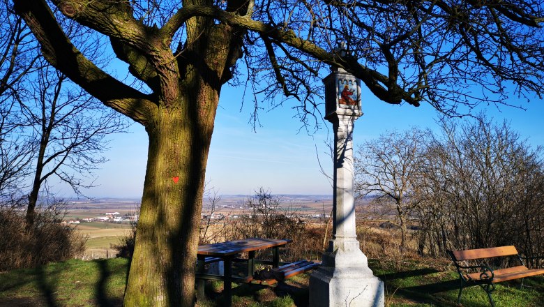 A large tree next to a stone wayside cross with benches and a sweeping view of the landscape.