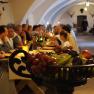 People at a knight's dinner in a vaulted hall with a bowl of fruit in the foreground.