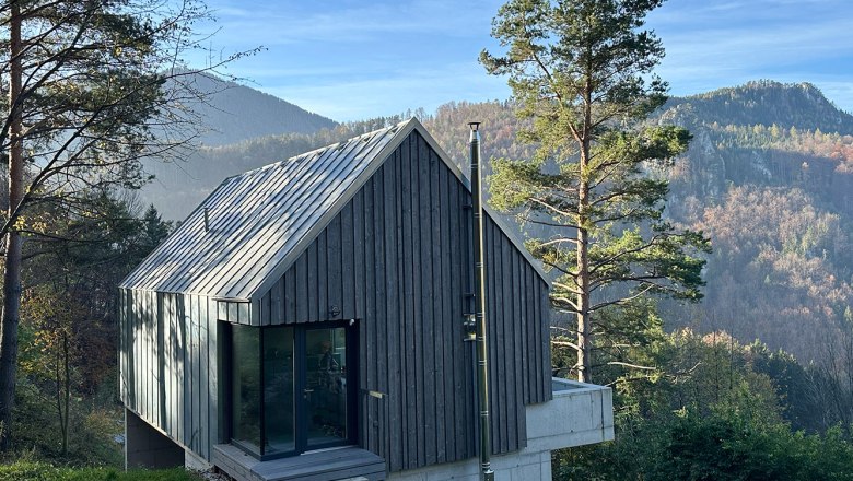 Modern wooden house in a mountainous landscape with trees and blue sky.