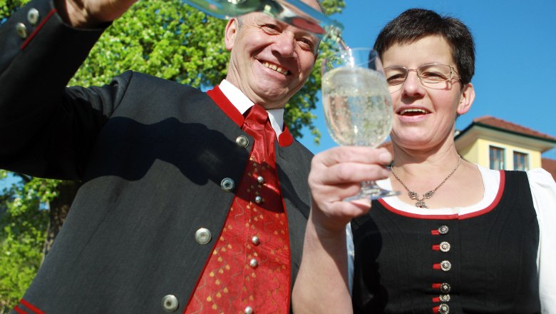 Two people in traditional dress clink glasses while a bottle is poured.