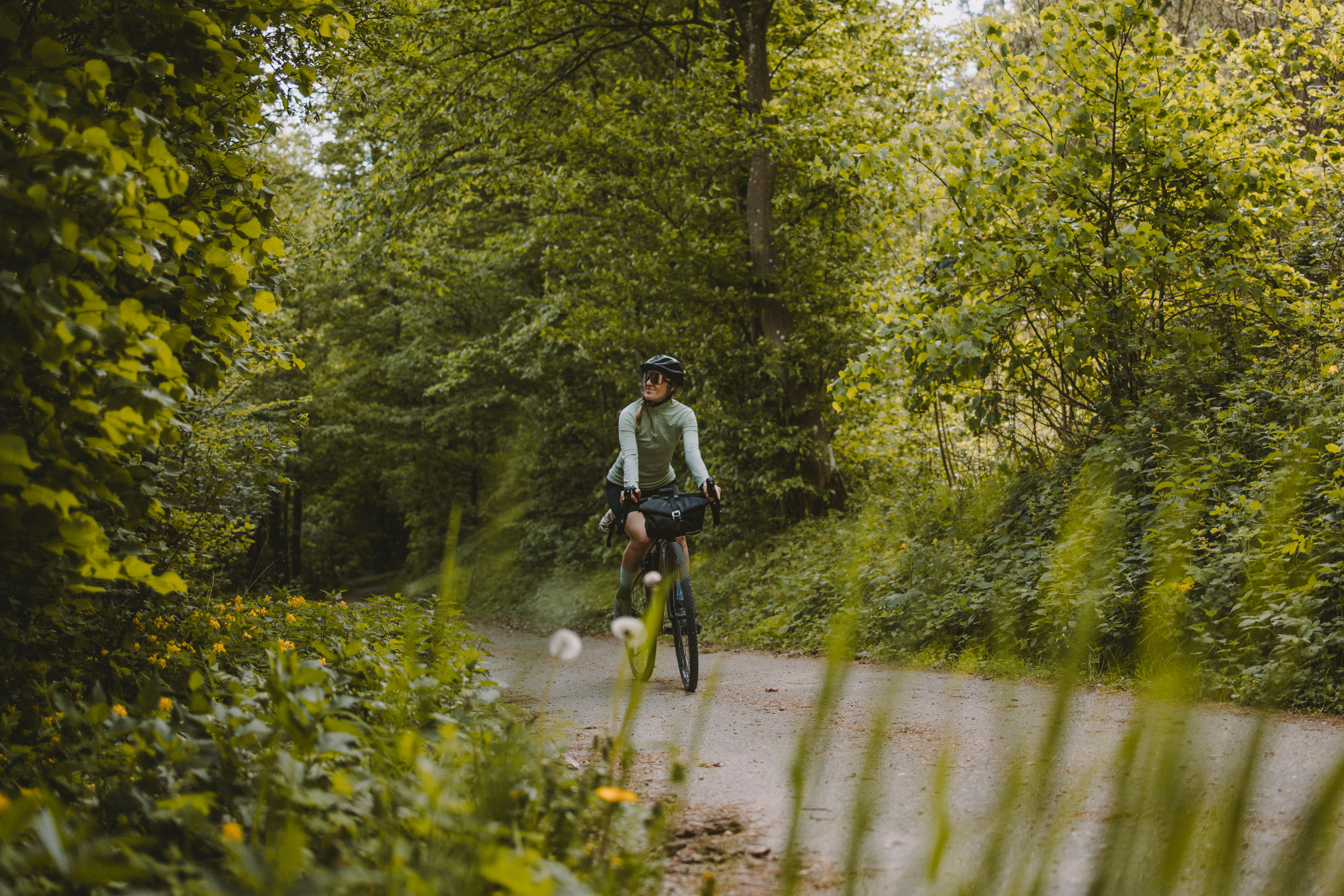 Ein Radfahrer genießt die frische Luft und die Ruhe der Natur auf einem malerischen Waldweg, umgeben von hohen Bäumen und sanften Hügeln. Die Sonne strahlt durch das Blätterdach und schafft eine einladende Atmosphäre für Abenteuerlustige und Naturliebhaber.