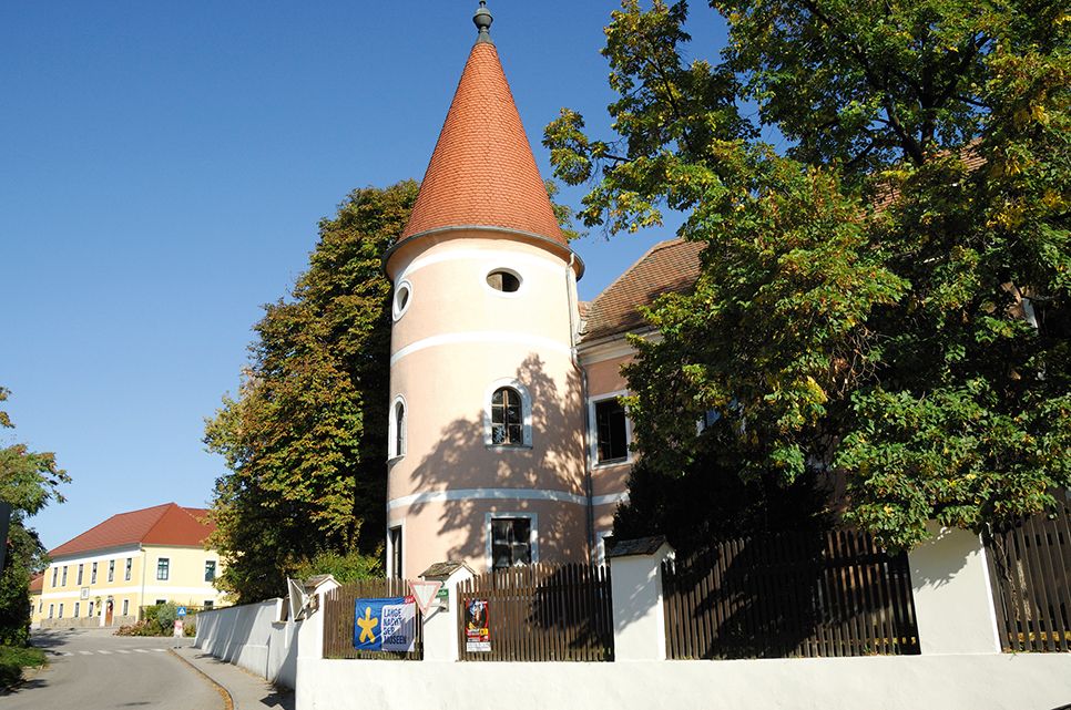 A pink building with a tower and red roof in Fels am Wagram, surrounded by trees and a blue sky.