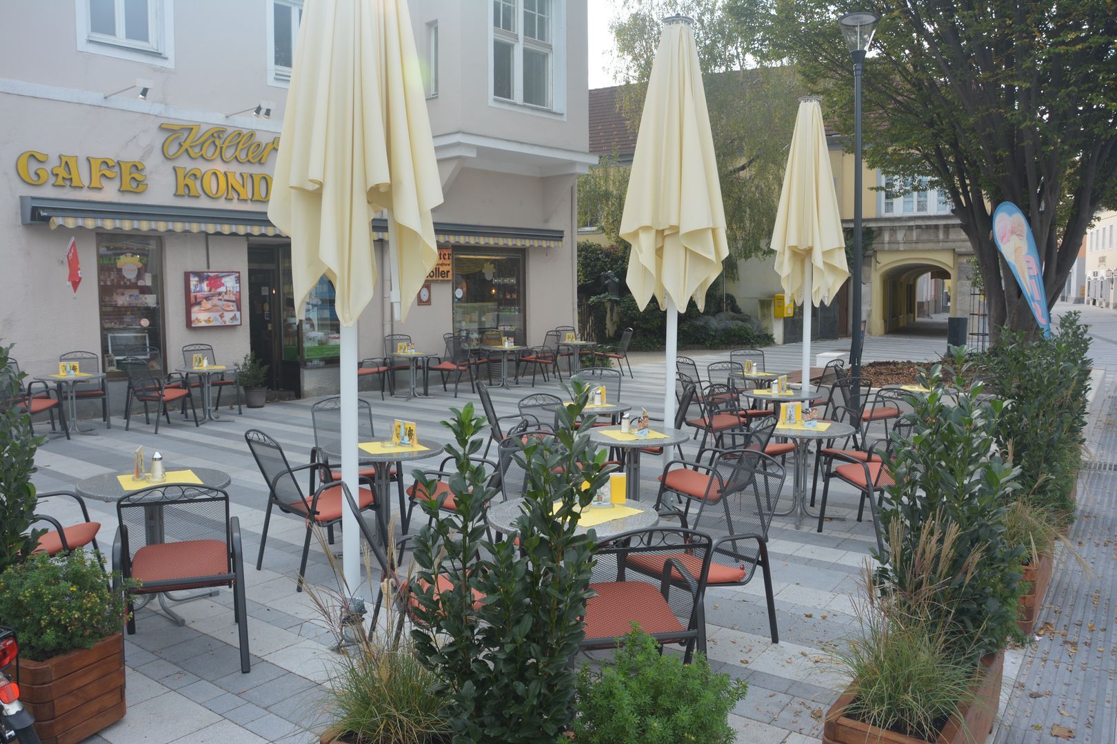 An empty pavement garden with tables and chairs in front of a café with yellow parasols.