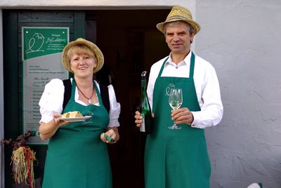 Two people in green aprons and straw hats hold wine and cake in front of a building.