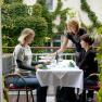 Two women sit at a table on a terrace while a waitress serves them food. A wall covered in ivy can be seen in the background.