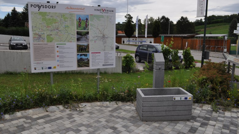 A drinking fountain next to an information board in Poysdorf, surrounded by flowers and parked cars.