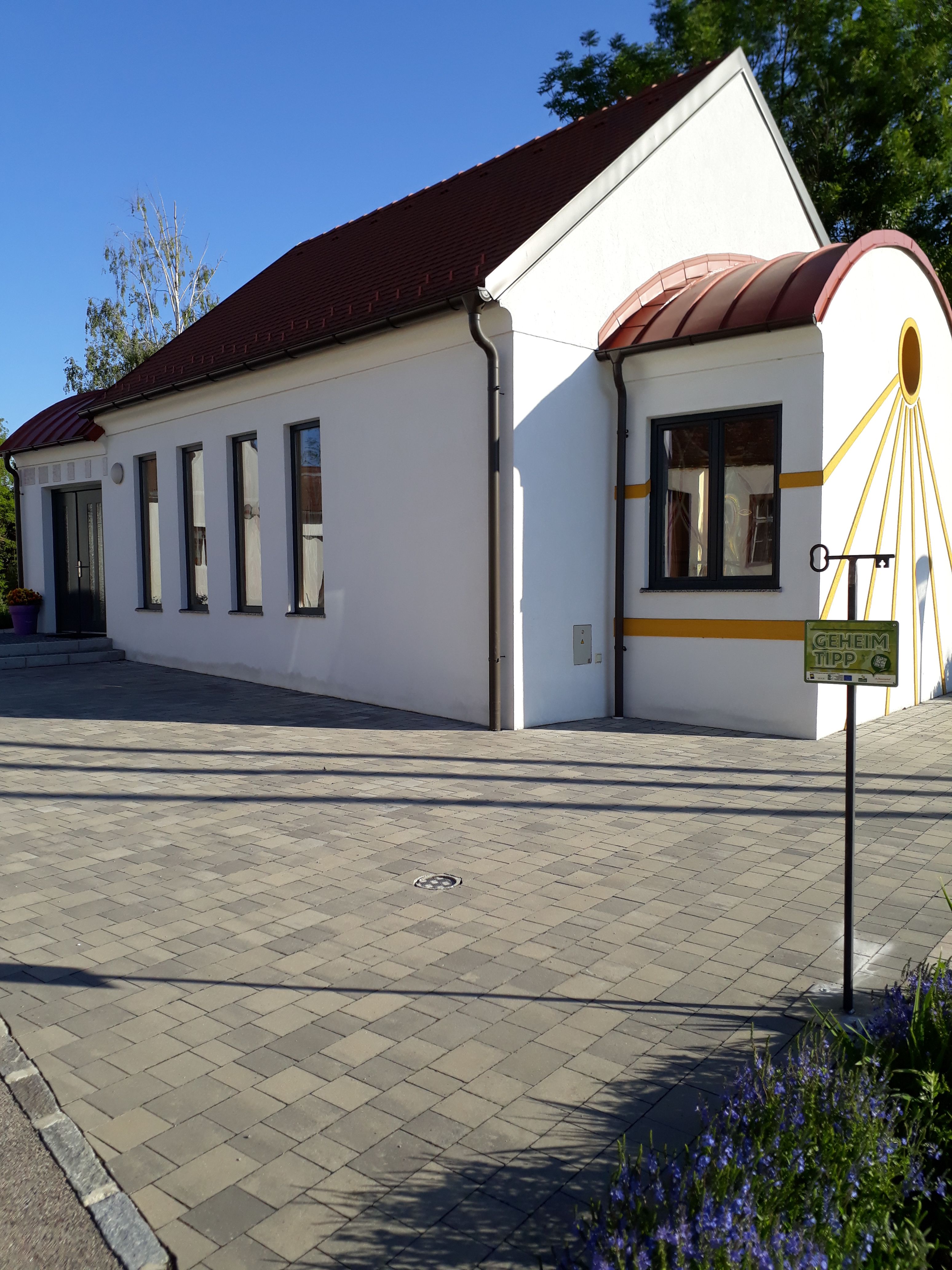 White chapel with red roof and yellow accents, surrounded by paved floor and plants.