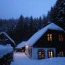 Snow-covered house in the forest at dusk with illuminated windows.